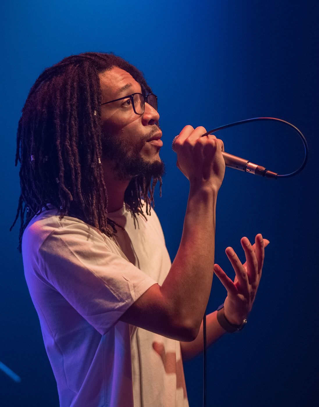 Singer with locs and glasses singing into a microphone under blue stage light