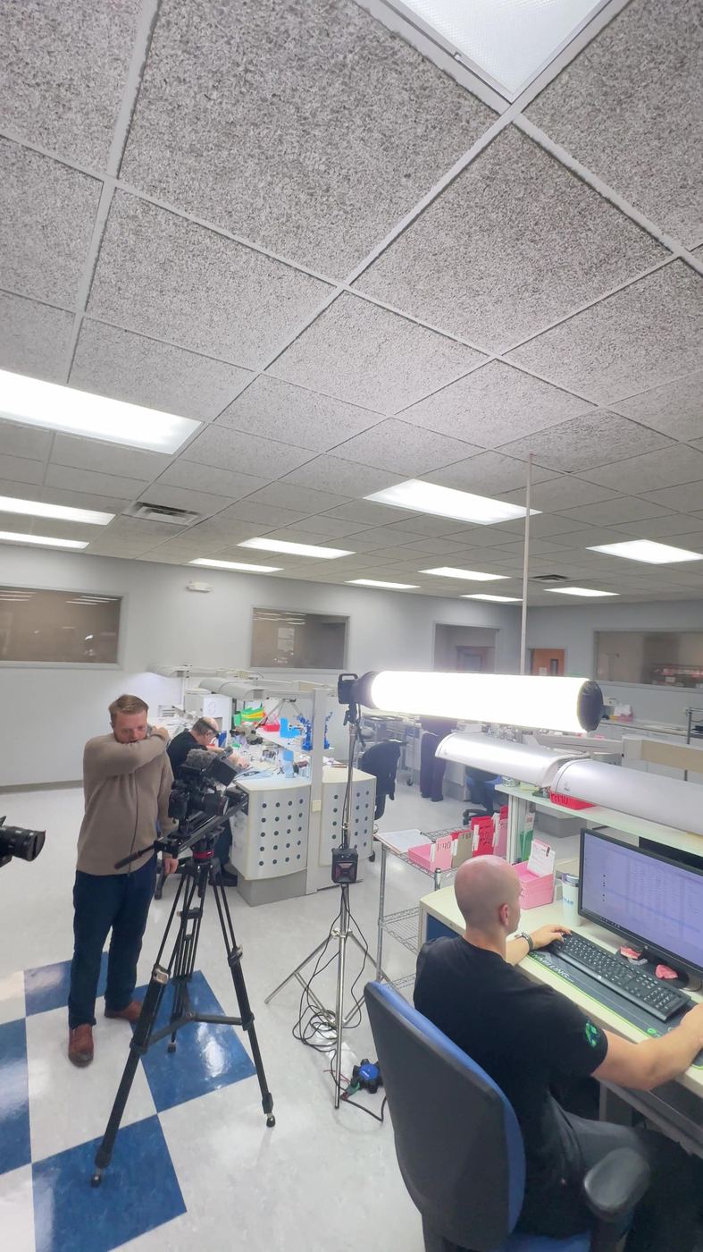 Camera operator filming a dental lab technician at his computer under a big light