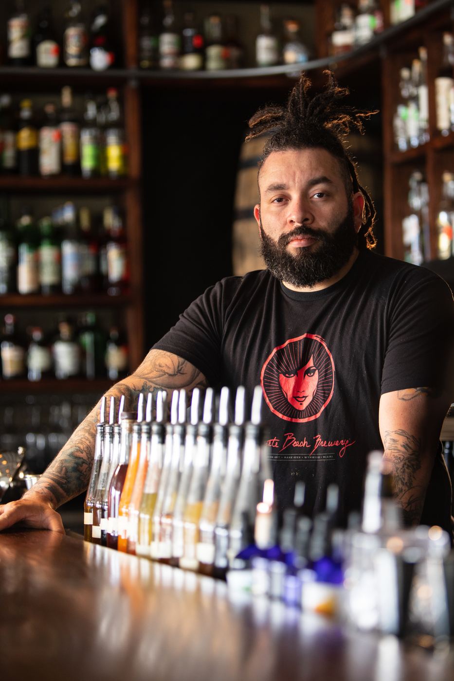 Bartender with a beard leaning on the bar in front of shelves of bottles