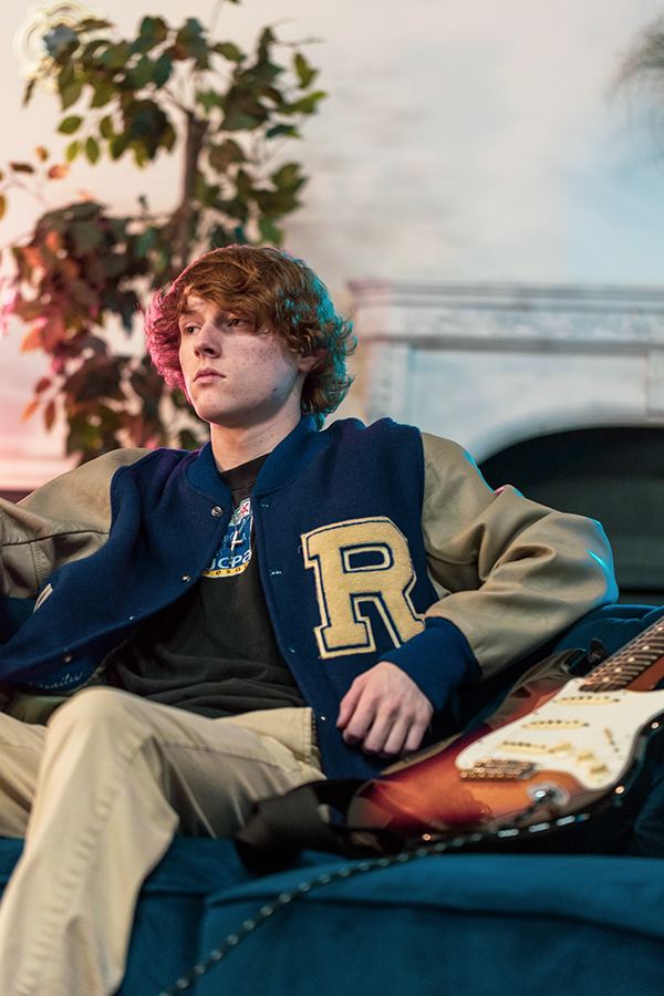 Young musician in a letterman jacket sitting on a couch with his guitar