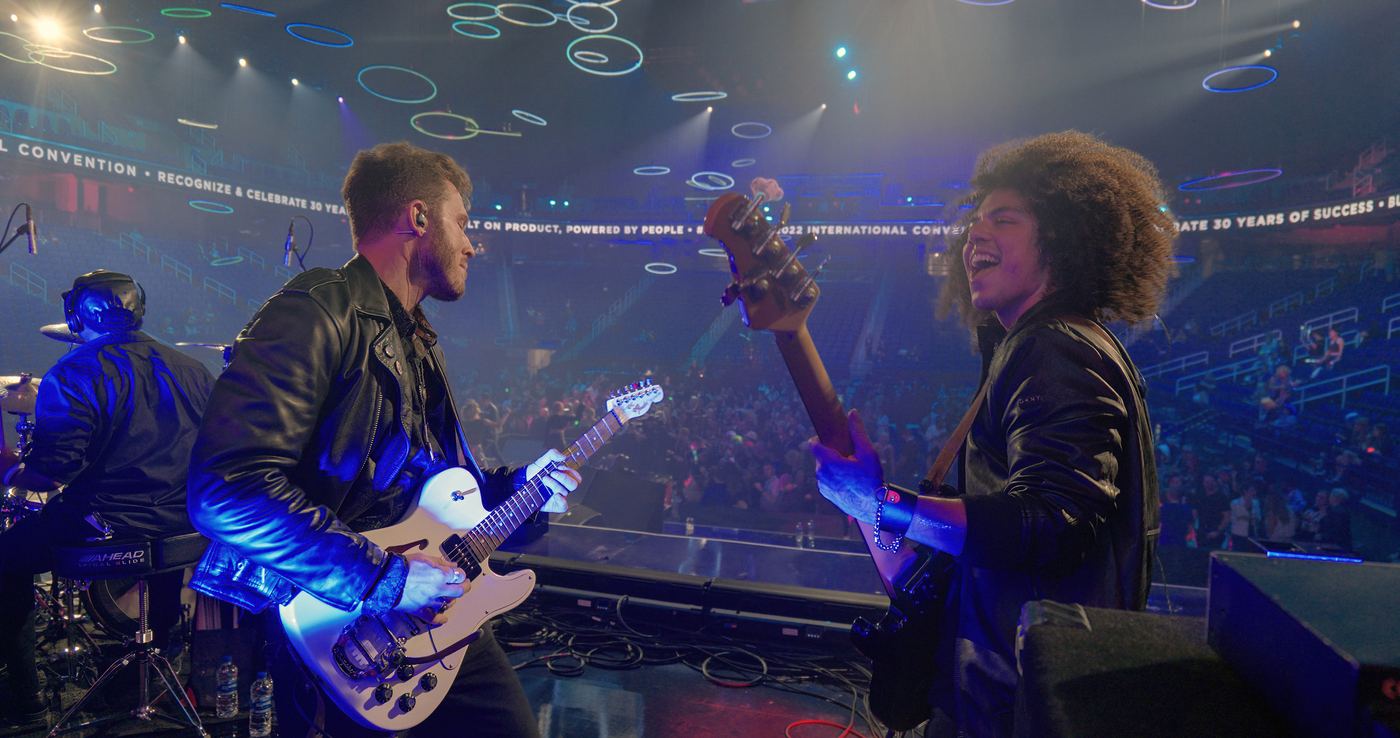 Two guitarists in leather jackets playing on a big convention stage with the crowd lit in blue