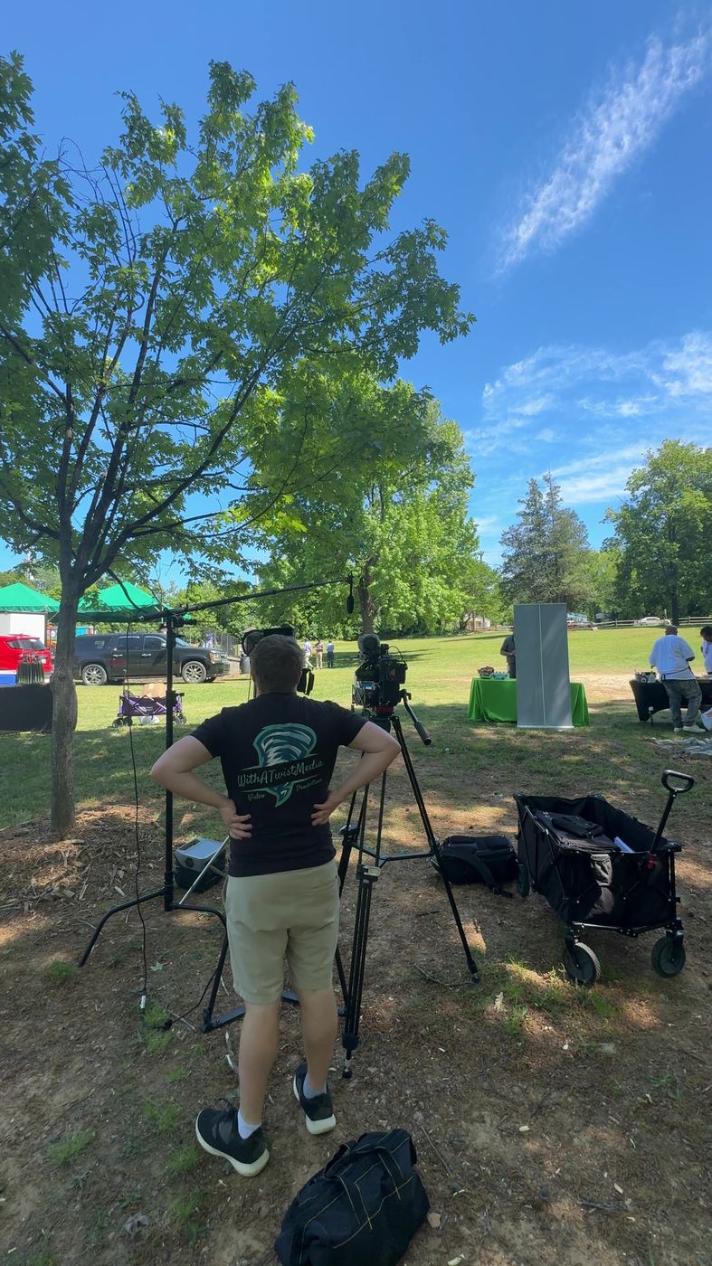 Crew in With A Twist shirts at two tripod cameras under a tree at an outdoor event