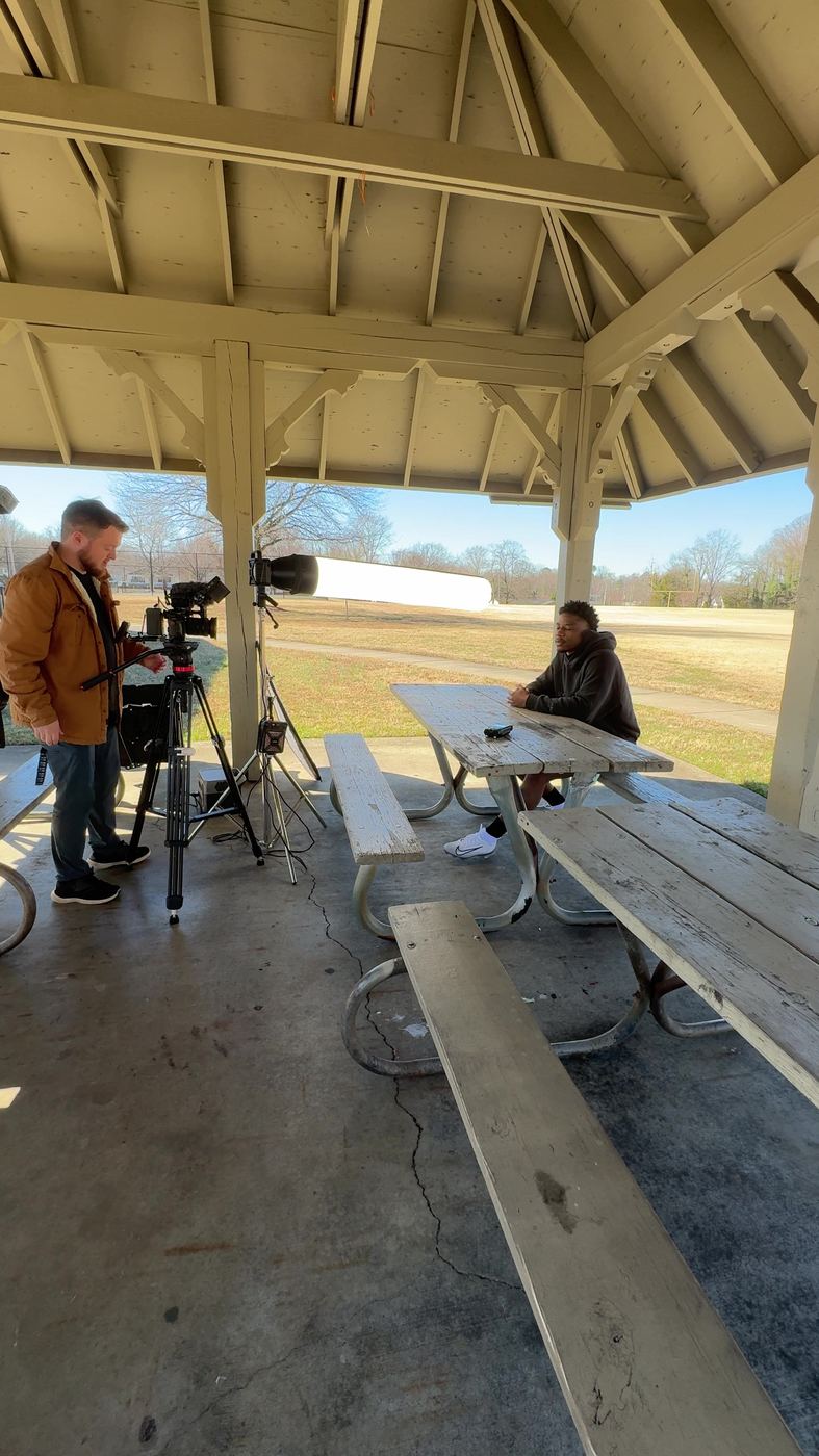 Outdoor interview under a park pavilion at a picnic table, crew at the camera