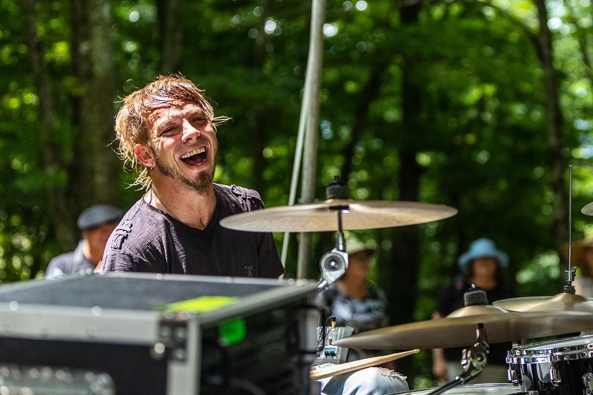 Drummer laughing behind his kit on an outdoor stage among the trees