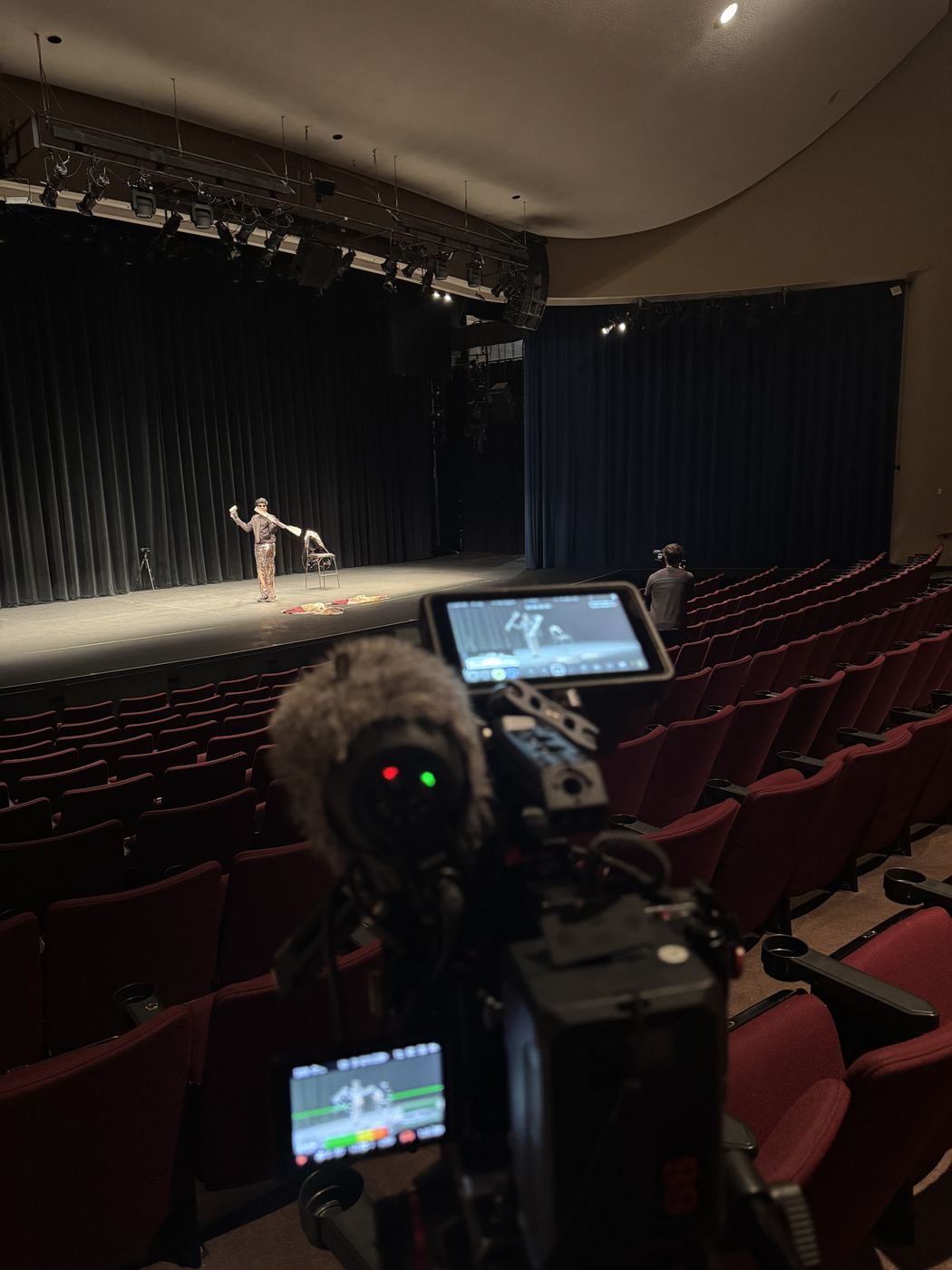 Camera with a fuzzy mic filming a performer on a theater stage from the seats