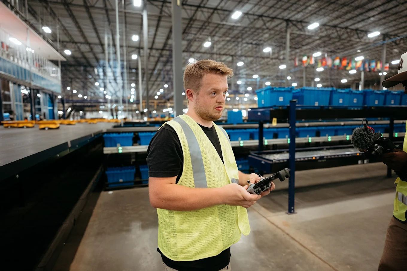 Dylan Smith in a safety vest holding a gimbal controller on a warehouse floor lined with blue bins