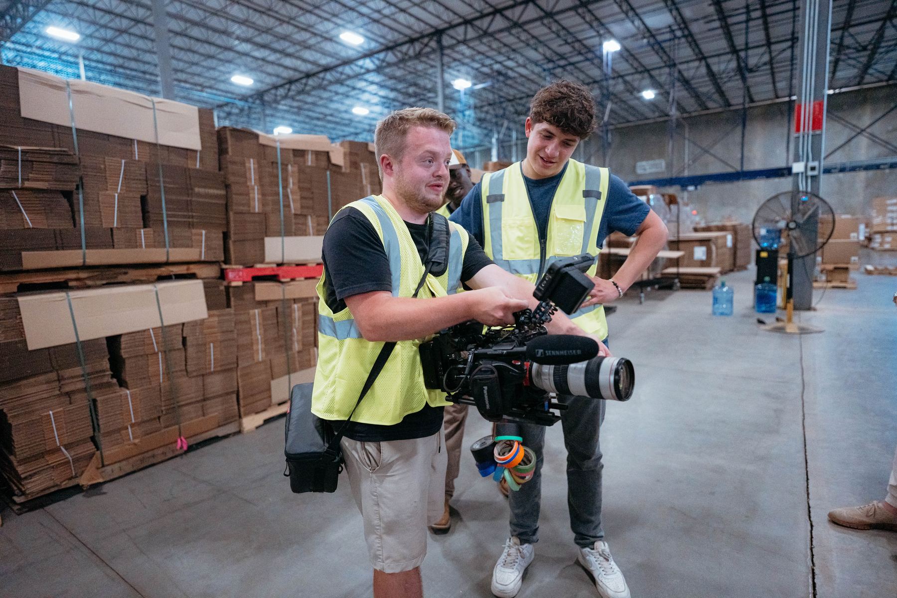 Dylan Smith and a crew member in safety vests checking a camera in a warehouse box aisle