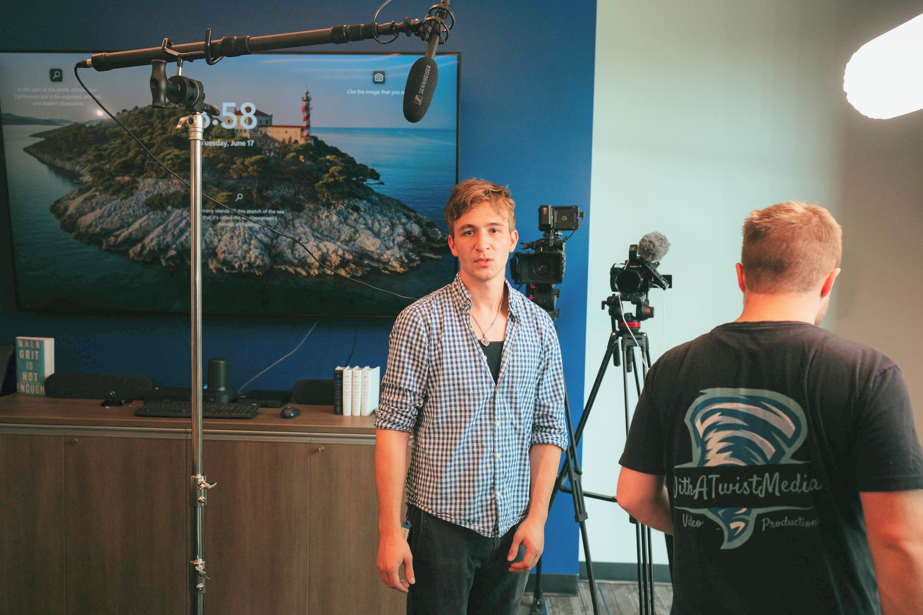 A man standing for an interview under a boom mic while a crew member checks the camera
